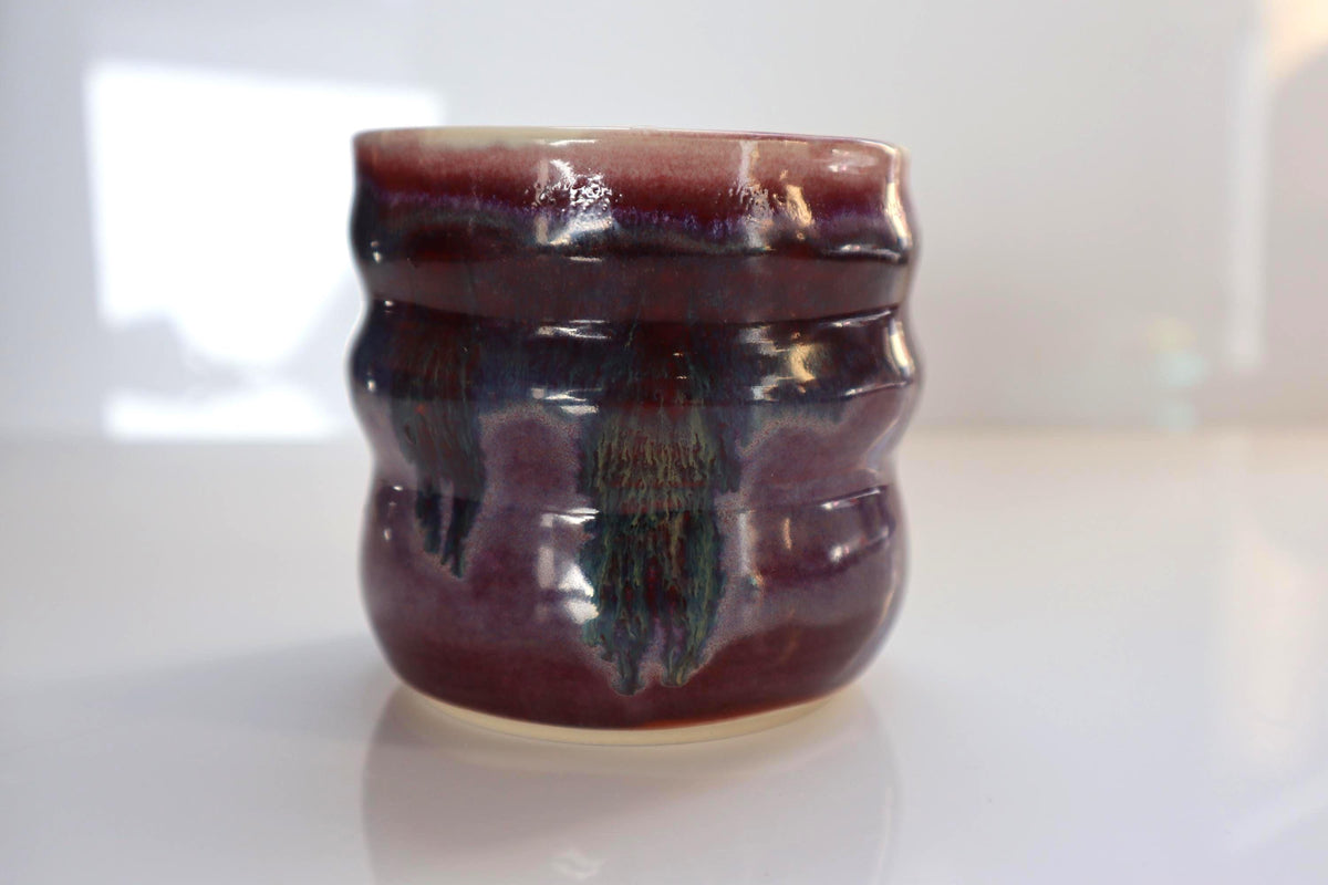 A small handmade ceramic cup with a wavy body and a glossy glaze, featuring a pink and purple color with green and blue drips, set against a bright white background.