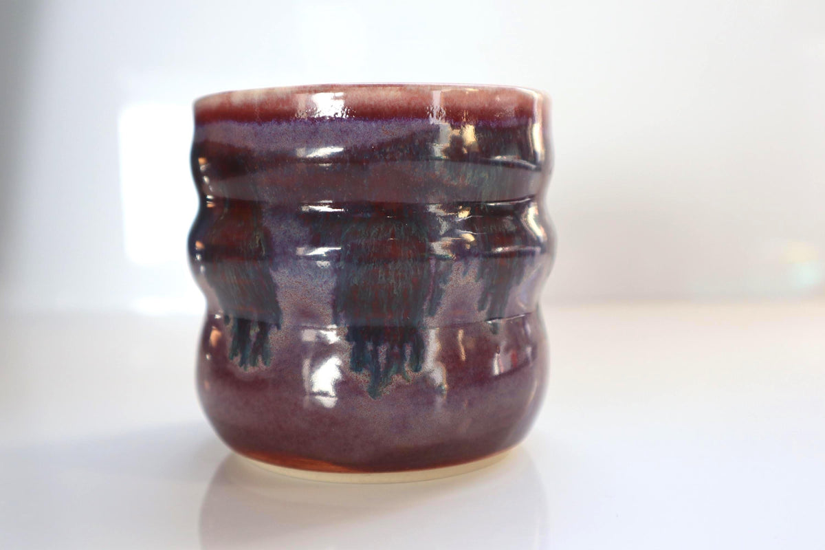 A handmade ceramic cup with a wavy, ribbed texture, featuring a glossy glaze of deep purple and pink, set against a bright white background.