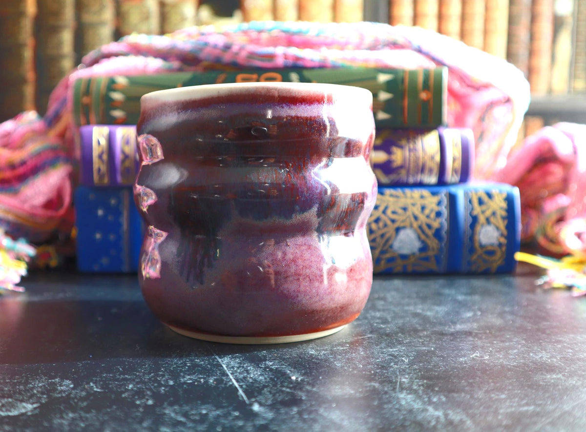 A handmade ceramic cup with a glossy,  purple and pink glaze, set on a dark tabletop with decorative books and colorful textiles in the background.