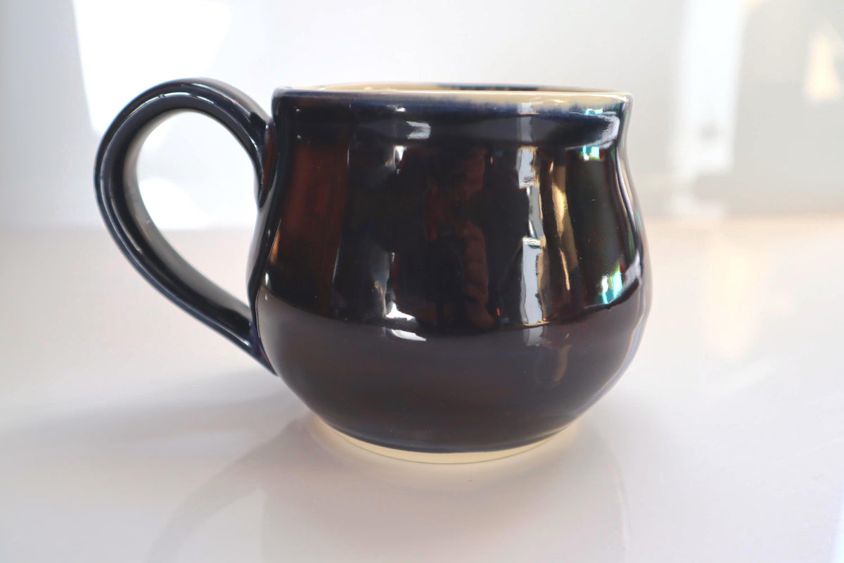 A glossy black ceramic mug with a curved handle, sitting on a reflective white surface.