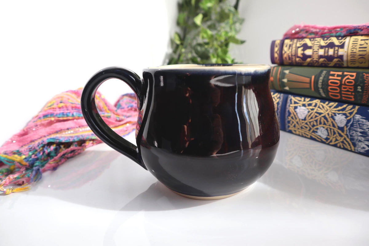 A glossy black ceramic mug with a curved handle, styled on a white surface with a colorful scarf, books, and a green plant in the background.