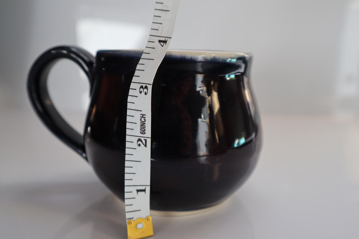 A glossy black ceramic mug with a handle, shown next to a white measuring tape to indicate its size, on a light gray background.