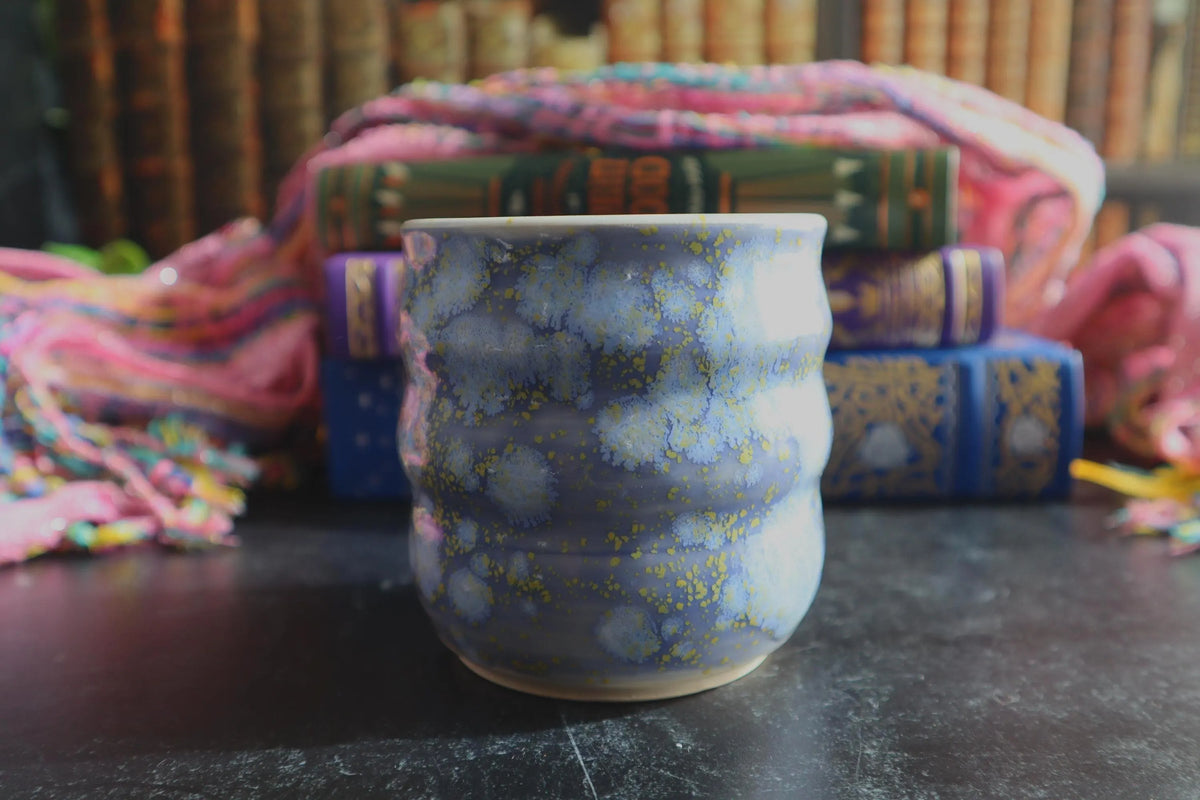 A handmade ceramic cup with a lavender glaze with yellow speckles, set against a background of colorful books and a pink scarf.