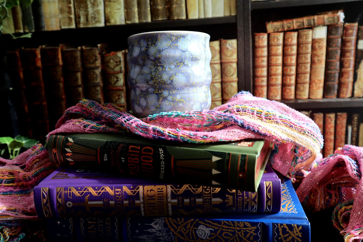 A decorative ceramic cup with a lavender, blue and yellow glaze sits on top of a stack of vintage hardcover books, draped with a pink and orange patterned knit scarf, set against a background of antique bookshelves.