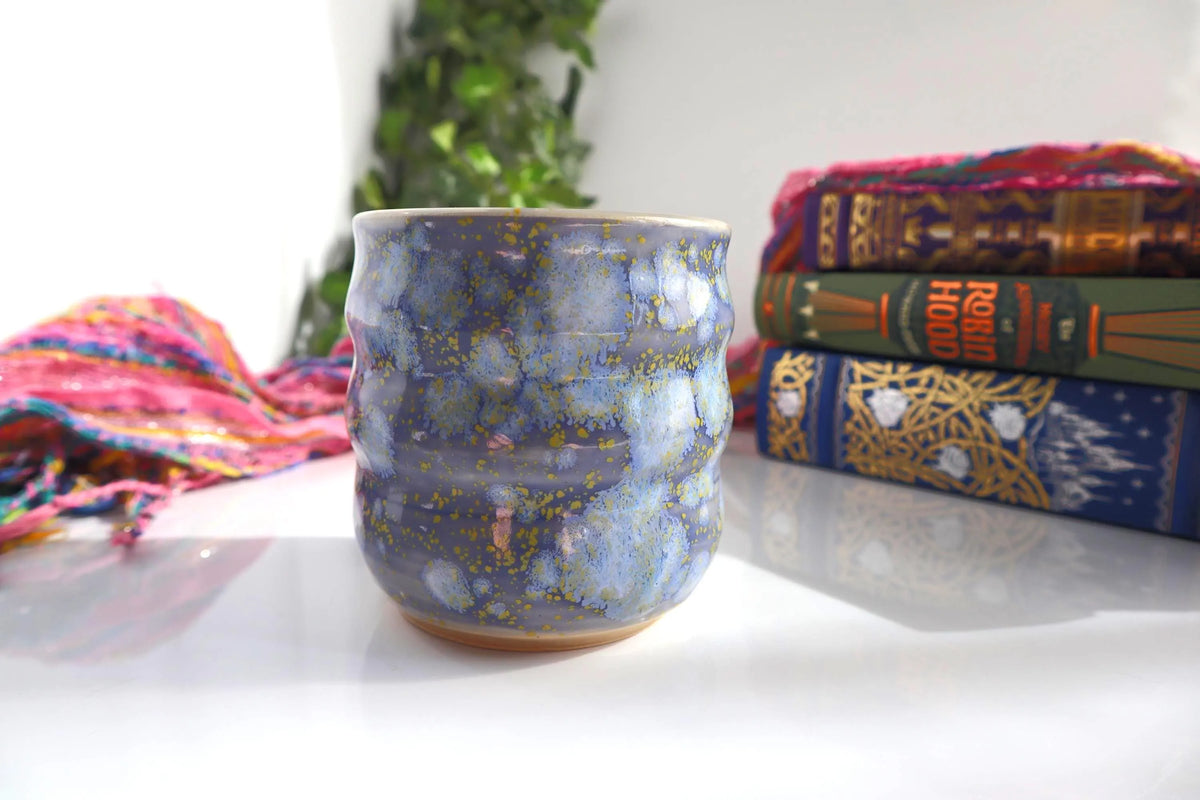 A handmade ceramic cup with a lavender and blue glaze styled on a white surface with books and a scarf in the background.