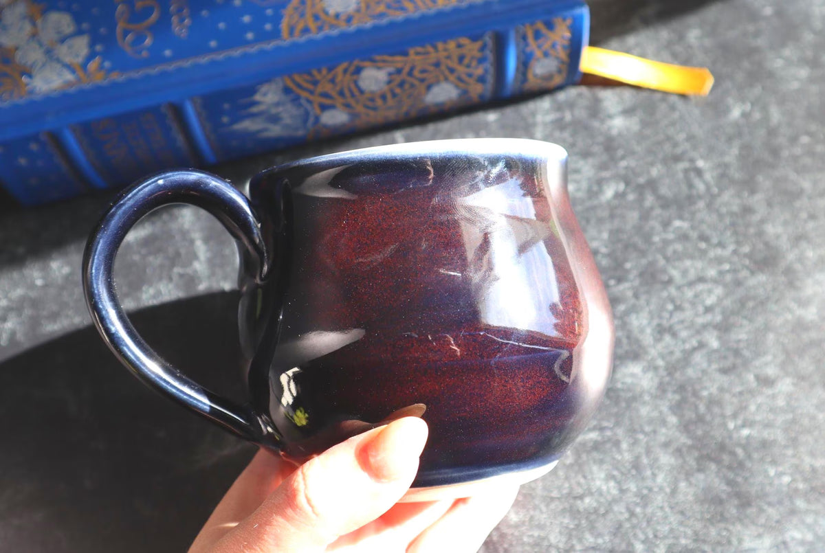 A hand holding a black and red ceramic mug with a glossy finish  with a blue patterned book in the background.