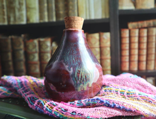 A small handmade ceramic bottle with a cork stopper, featuring a glossy dark pink and purple glaze with iridescent blue-green swirls, resting on a pink woven scarf in front of a blurred background of bookshelves.